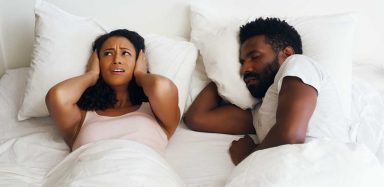 A man snoring in bed while a woman puts a pillow over her ears.