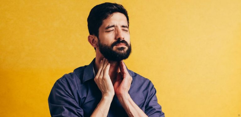 A man in a blue shirt standing in front of a yellow background. He's holding his hands to his neck, as if experiencing a sore throat.